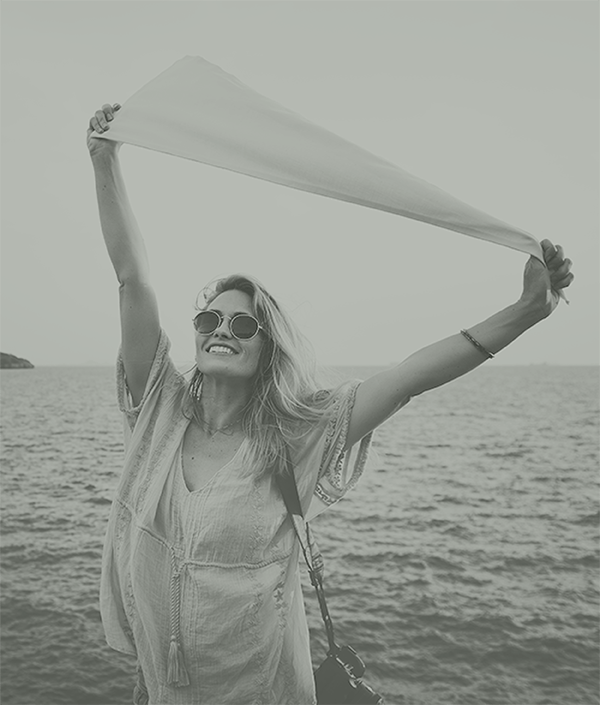 Woman celebrating feeling independent from mental health issues.  She's holding a scarf above her head in front of an ocean smiling and wearing sunglasses